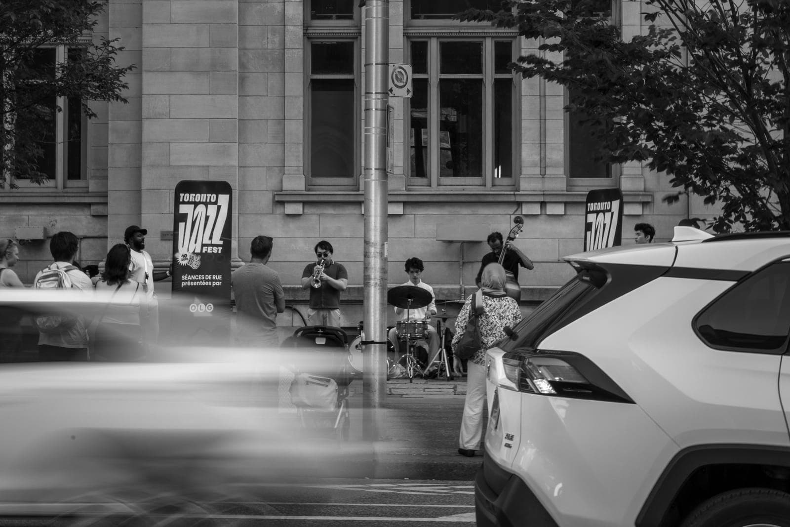Joseph Funk's trio plays trumpet, drums, and upright bass on the sidewalk between Toronto Jazz Festival banners as a car blurs past