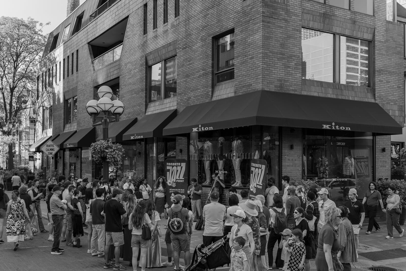 A large crowd packs the Yorkville sidewalk outside the Kiton storefront to watch Joseph Funk's trumpet-and-upright-bass duo play beside a Toronto Jazz Fest Sidewalk Sessions banner