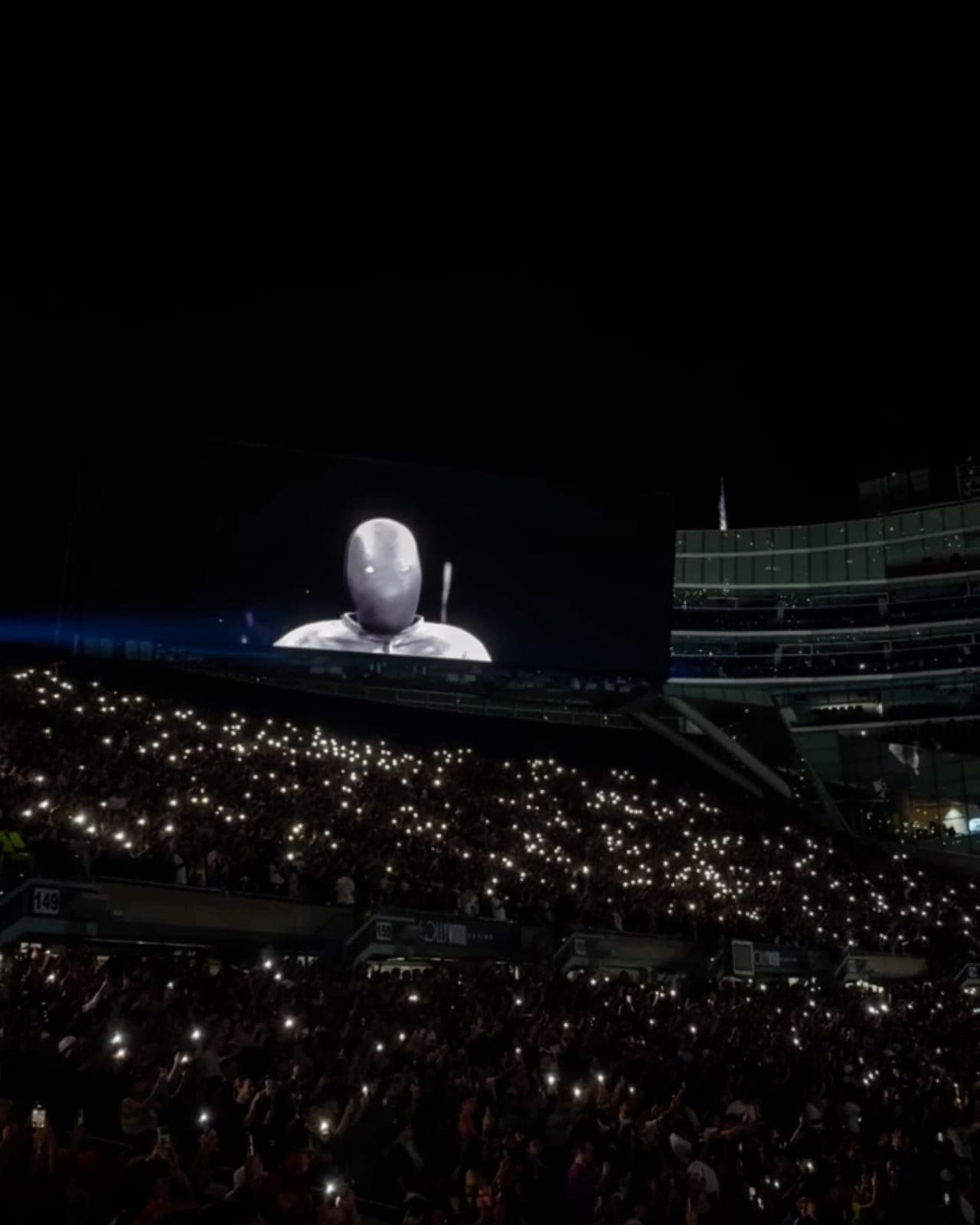 The Soldier Field jumbotron shows Ye in a full-face silver mask and hood while the stands below fill with phone flashlights
