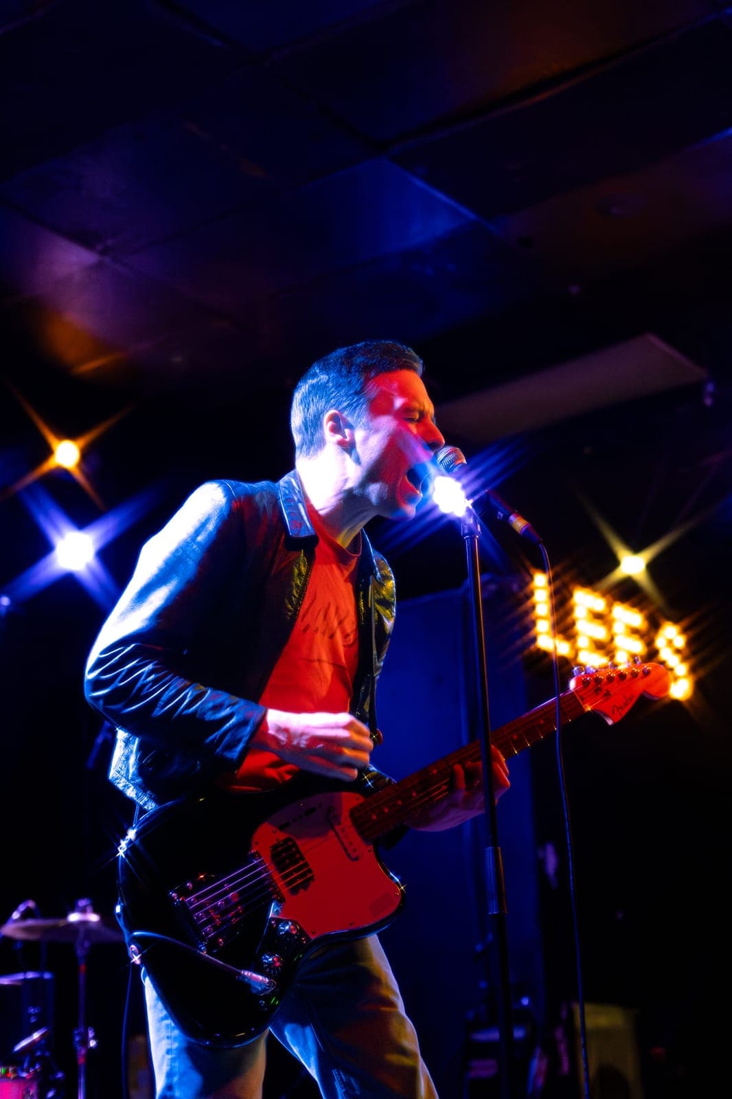 The Cribs' Ryan Jarman belts into the mic mid-song with a red Fender guitar slung low, the marquee 'LEES' sign glowing behind him at Lee's Palace in Toronto
