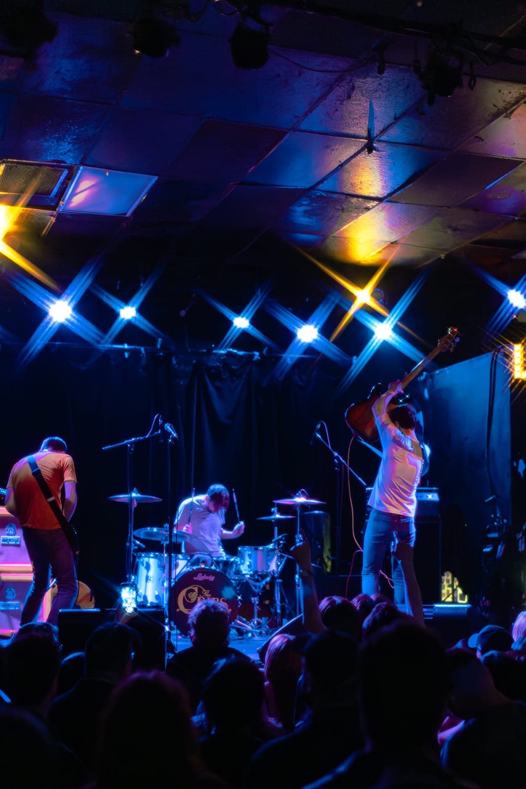 View from the crowd as The Cribs' Gary Jarman raises his bass overhead, with Ross Jarman on drums and Ryan Jarman on guitar framed by blue X-shaped stage lights at Lee's Palace in Toronto