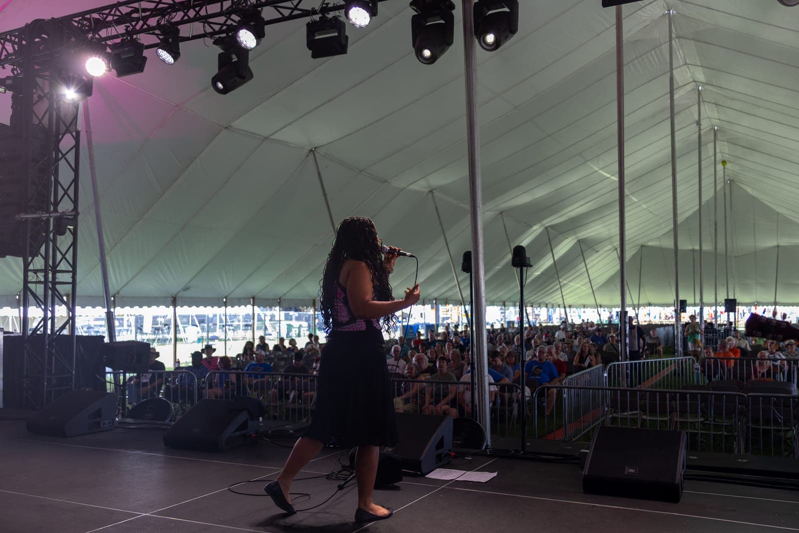 Isy Aboagye sings into a handheld mic on the darkened festival stage at Kitchener Blues Festival, silhouetted against the seated crowd under the big-top tent