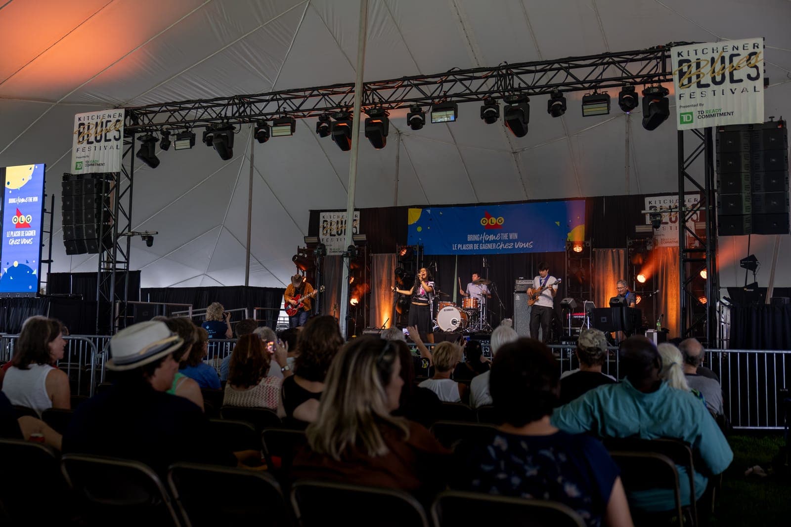 A wide view of Isy Aboagye and her band performing on the Kitchener Blues Festival stage beneath the festival banners, the seated crowd watching from folding chairs