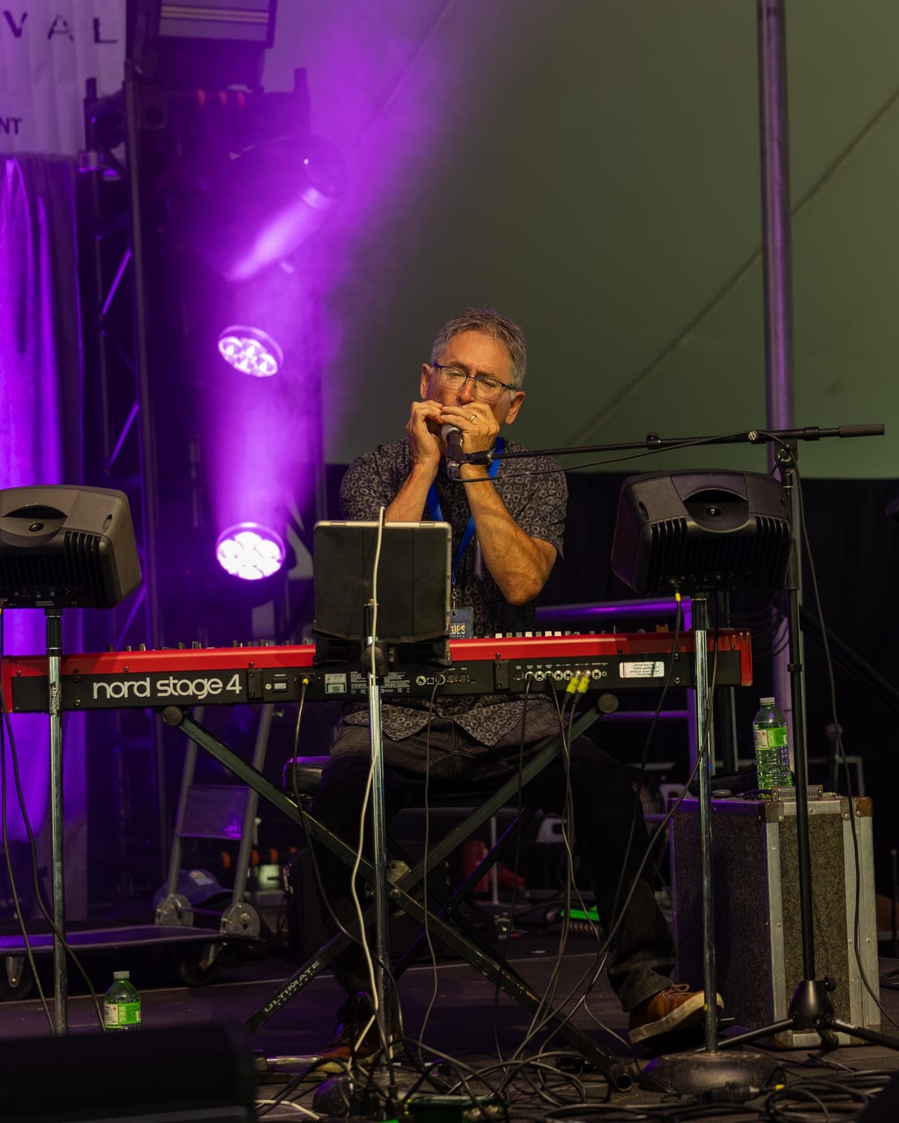 Isy Aboagye's keyboardist cups a harmonica to the mic over his red Nord Stage 4 under a purple spotlight at Kitchener Blues Festival