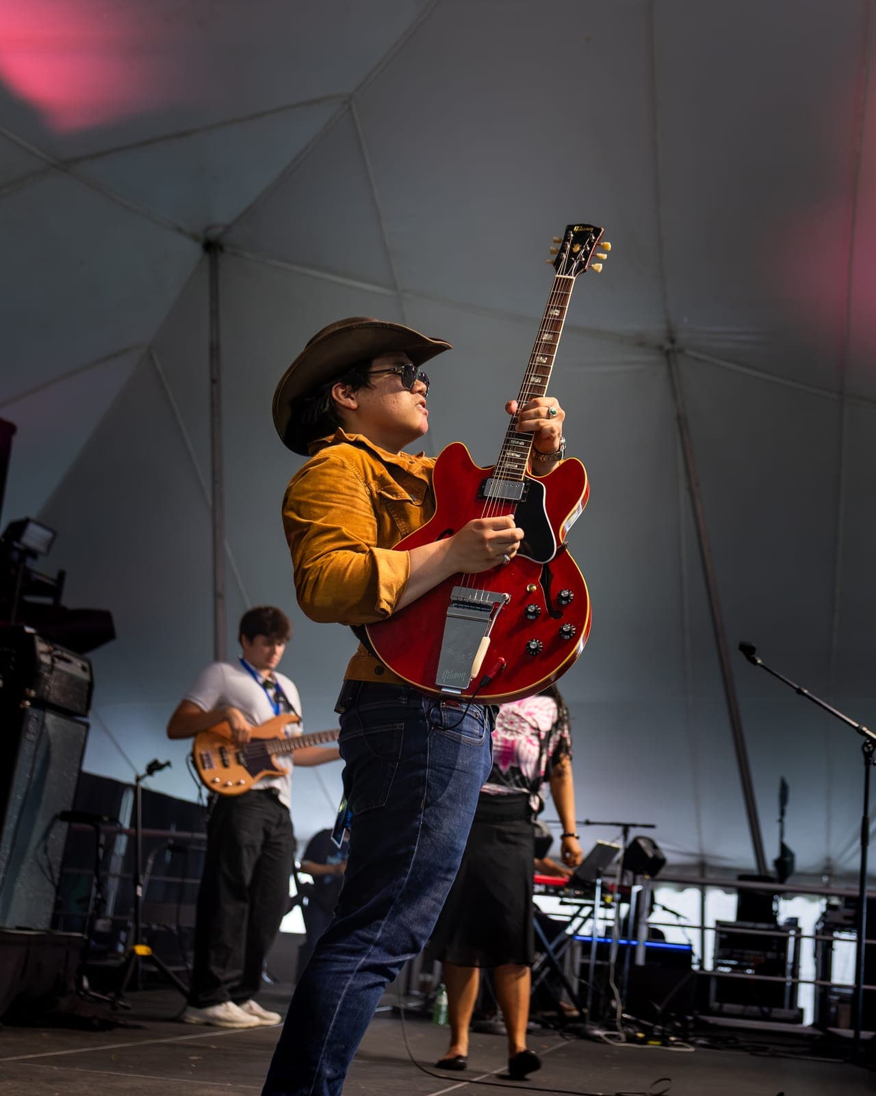 Isy Aboagye's guitarist thrusts his red Gibson semi-hollow into the air in a mid-song action shot at Kitchener Blues Festival, cowboy hat and mustard jacket catching the stage light