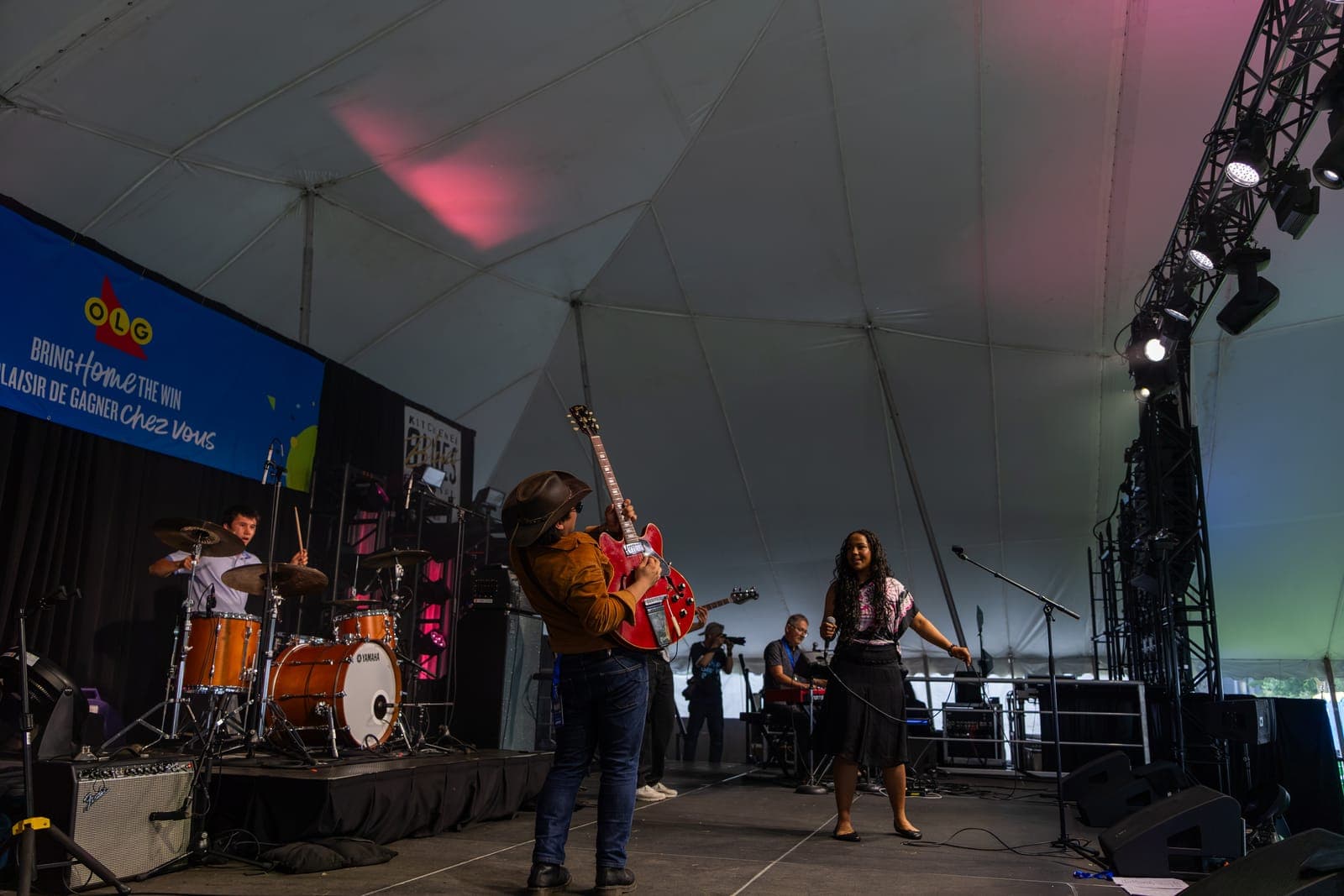 A wide view across the Kitchener Blues Festival stage as Isy Aboagye's guitarist hoists his red Gibson toward her while she sings, the drummer at his Yamaha kit on the left and the keyboardist behind