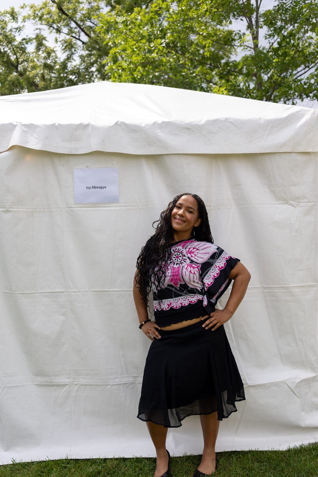 Isy Aboagye poses with a hand on her hip outside her green-room tent at Kitchener Blues Festival, her name taped to the white canvas beside her and summer trees overhead
