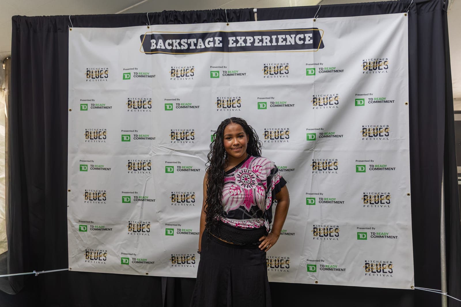 Isy Aboagye stands alone in front of the 'Backstage Experience' step-and-repeat banner at Kitchener Blues Festival, smiling with one hand resting on her hip