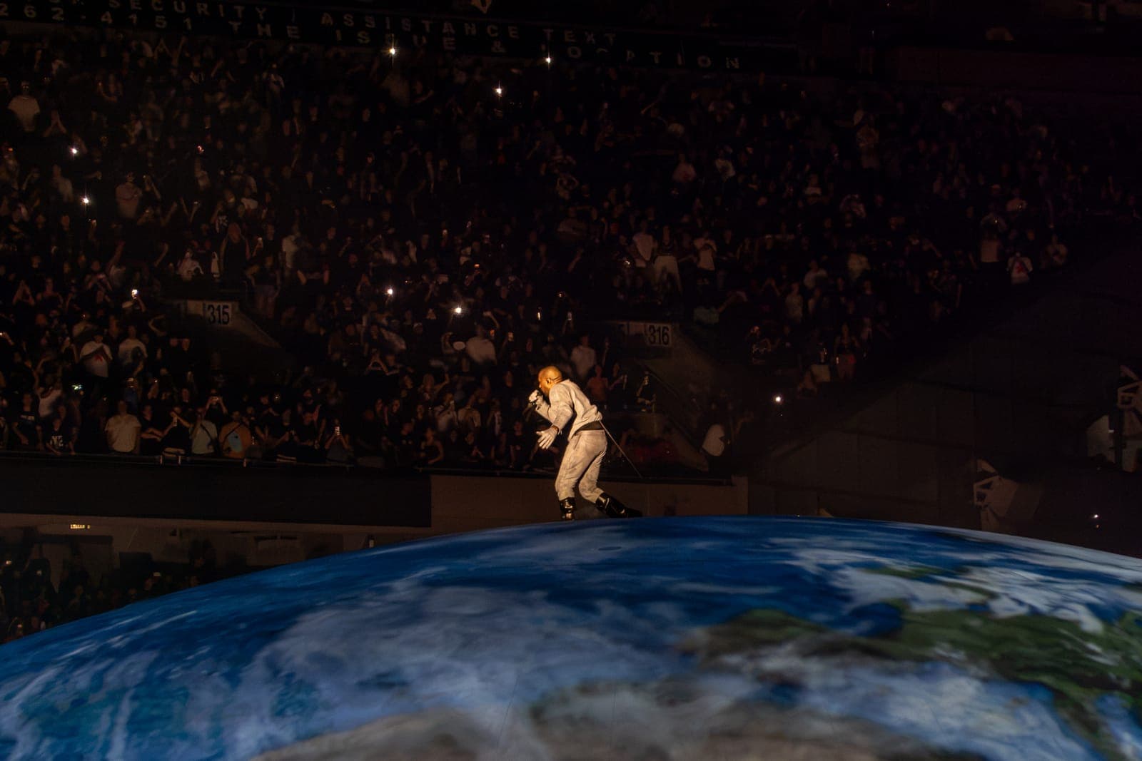 Ye crouches forward into the mic in a pale grey jumpsuit, the packed upper tiers of Soldier Field rising behind him and the projected Earth curving away below