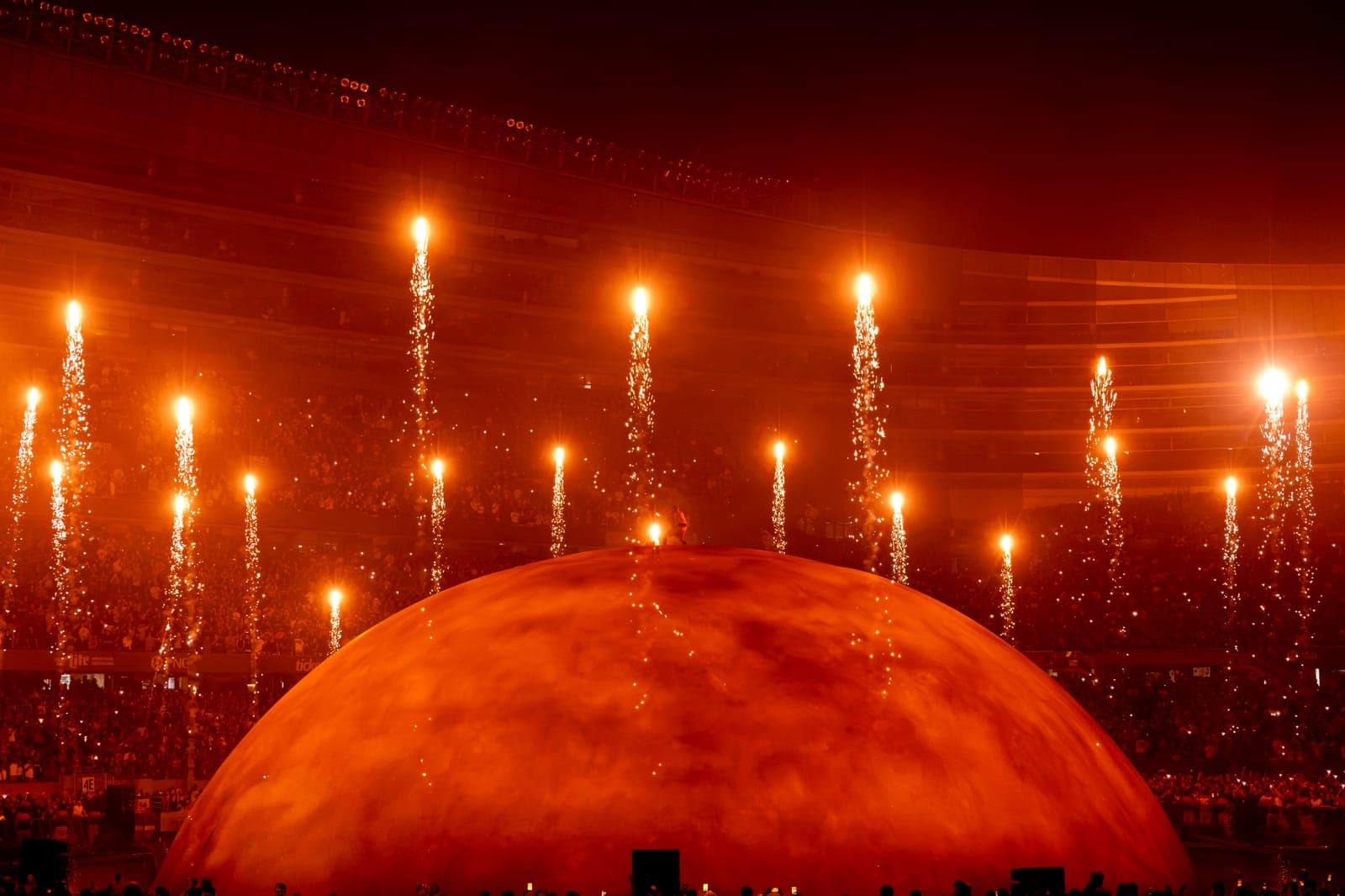 The stage dome burns deep orange as fireworks shoot upward around it, a lone figure silhouetted at the summit