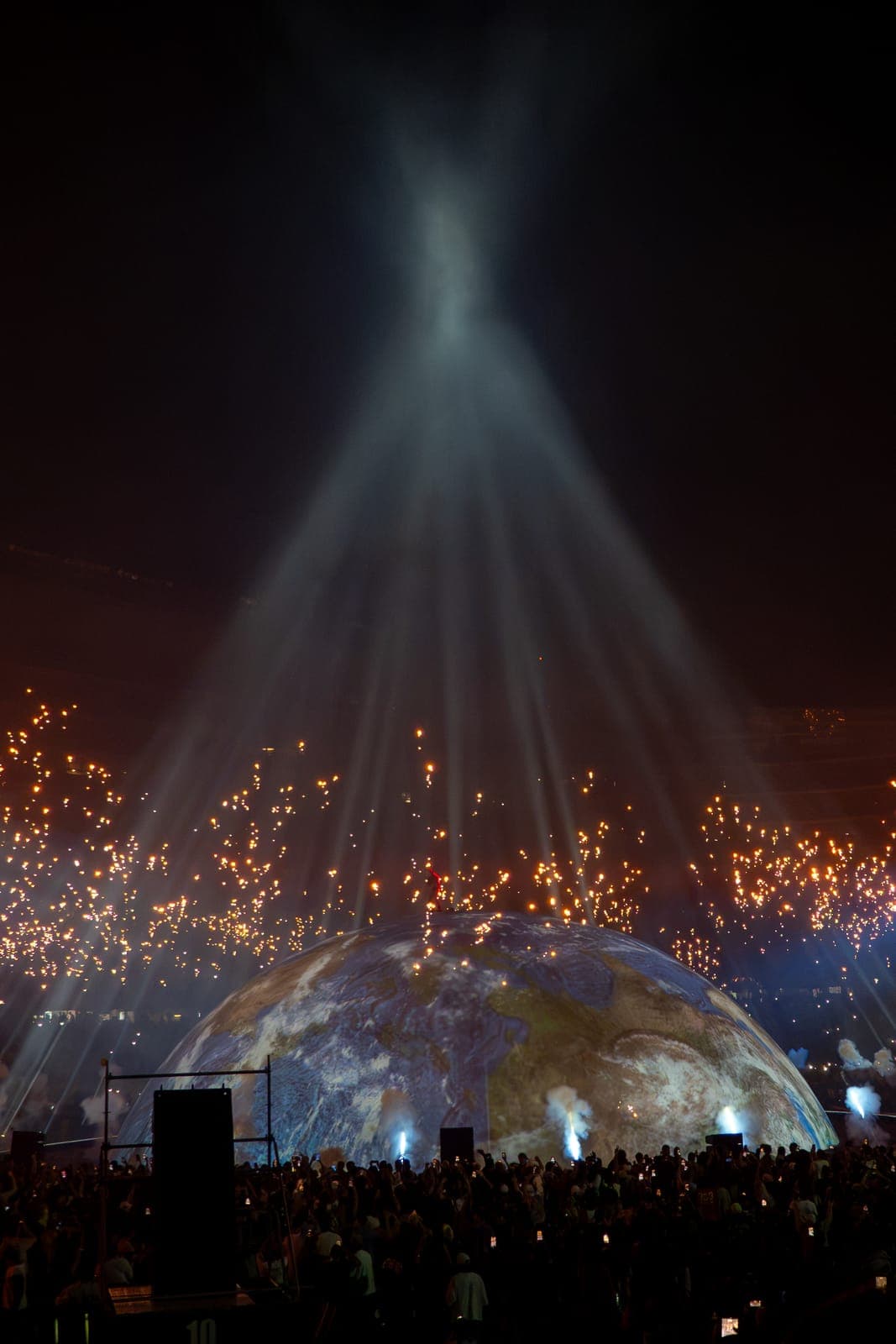 Shafts of white light pour down onto the projected Earth as orange sparks arc overhead and the crowd watches from the floor