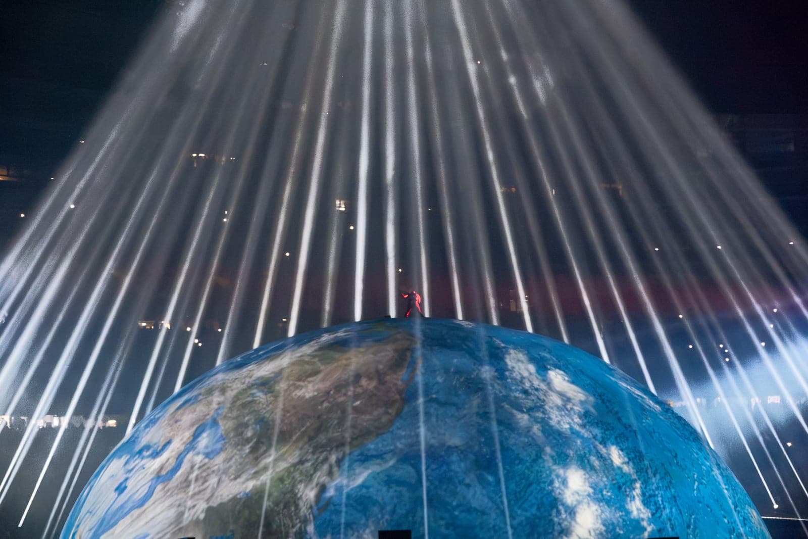 A curtain of white beams falls in parallel lines onto the blue Earth projection, a small red-lit figure standing at its crown