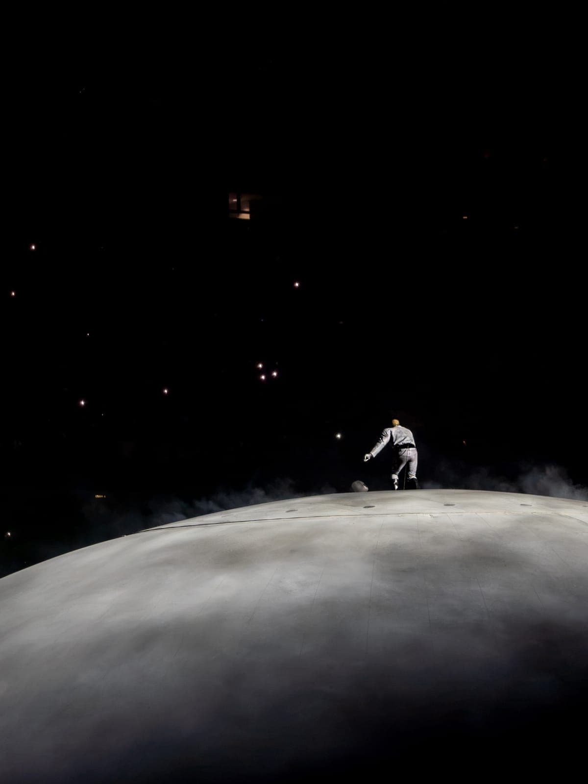 Ye in pale grey walks the curve of the unlit dome through drifting haze, the stadium almost entirely black around him