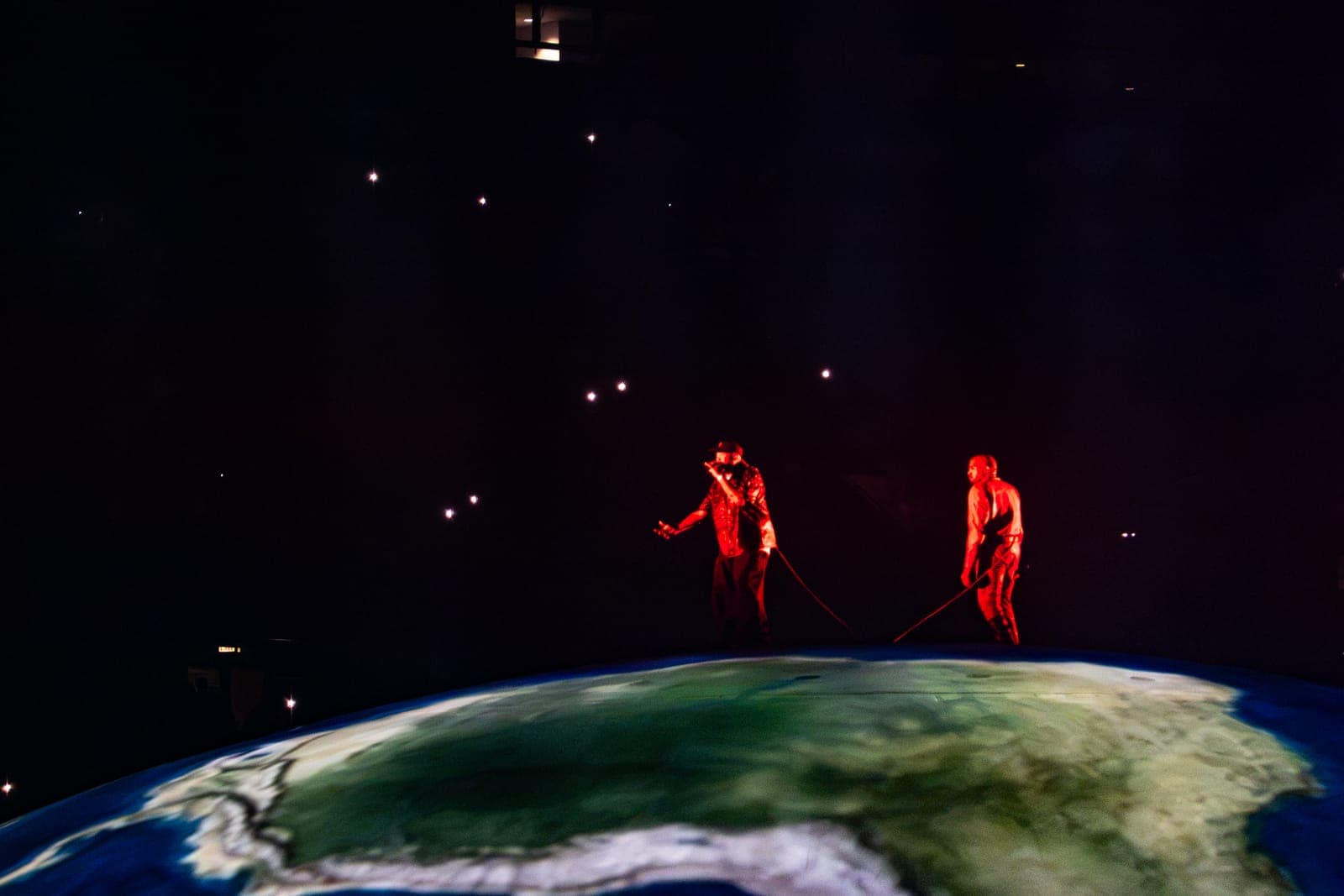 Twista leans back with arms outstretched beside Ye on the projected Earth, both washed in red under a starfield of lights