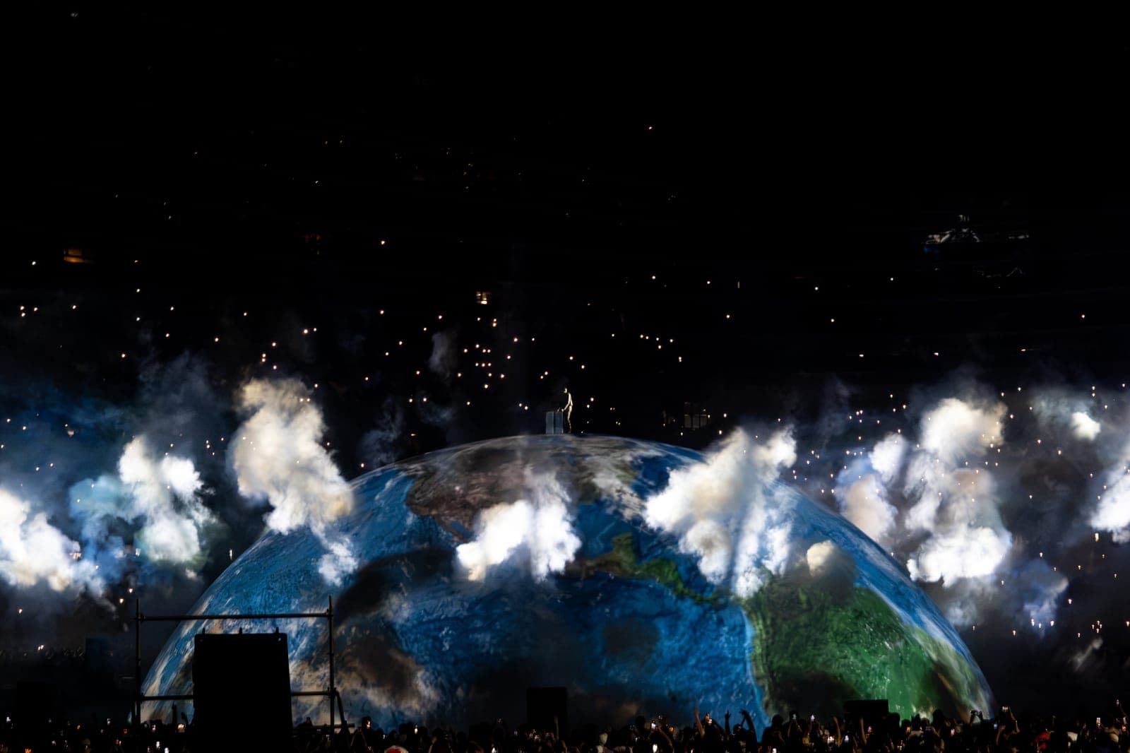 Ye stands alone on top of the giant projected Earth at Soldier Field as plumes of smoke burst around the globe and thousands of phone lights fill the dark stands