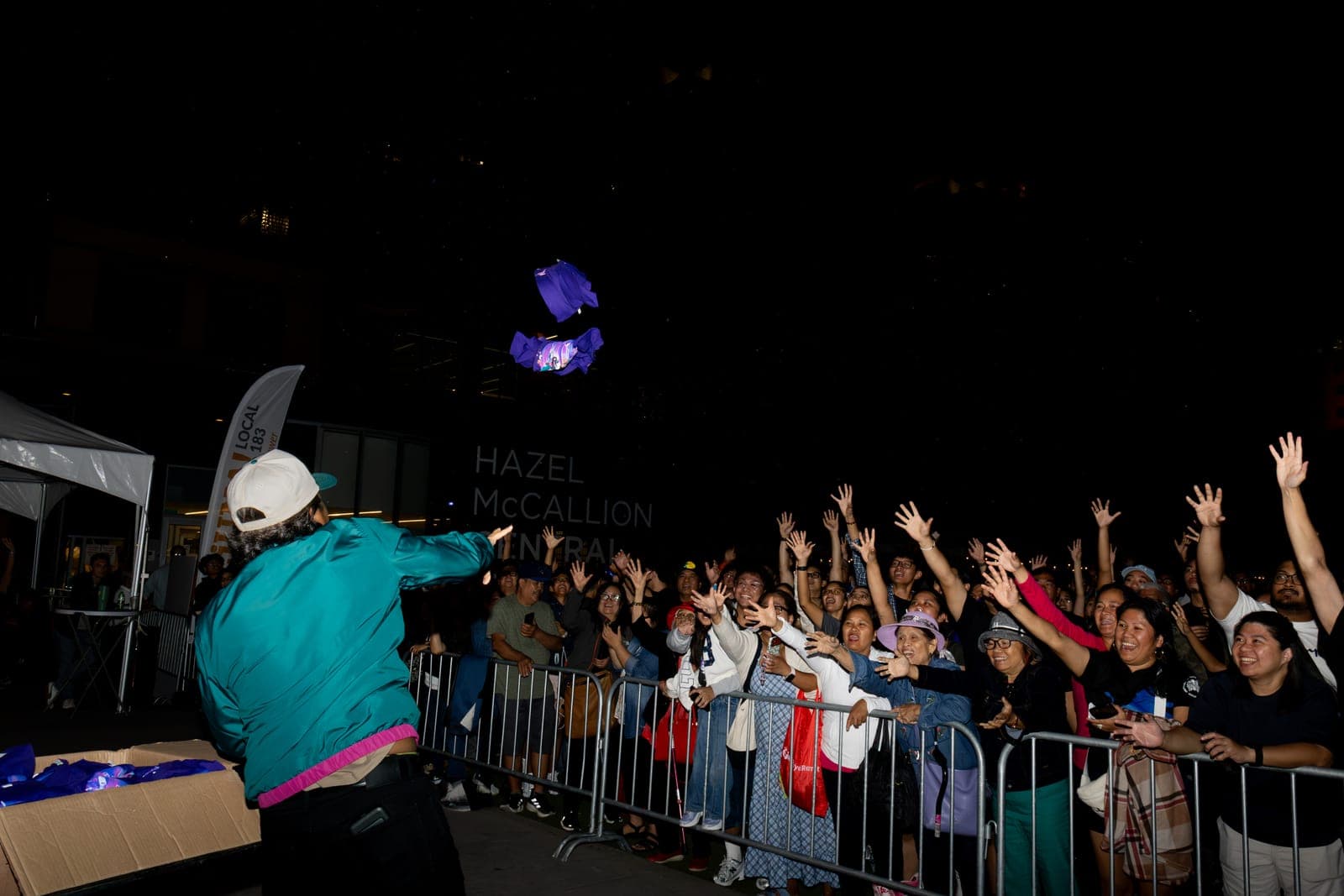A crew member in a teal jacket launches purple t-shirts into a crowd of raised hands at Mississauga Celebration Square