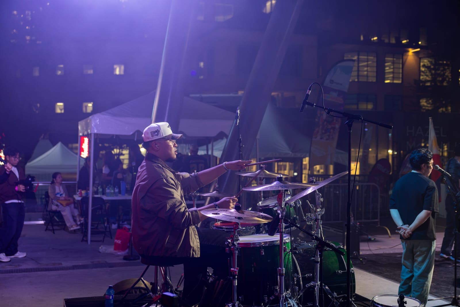 AJ Rafael's drummer points a stick mid-fill behind his green kit on the outdoor stage at Mississauga Celebration Square, festival tents glowing behind