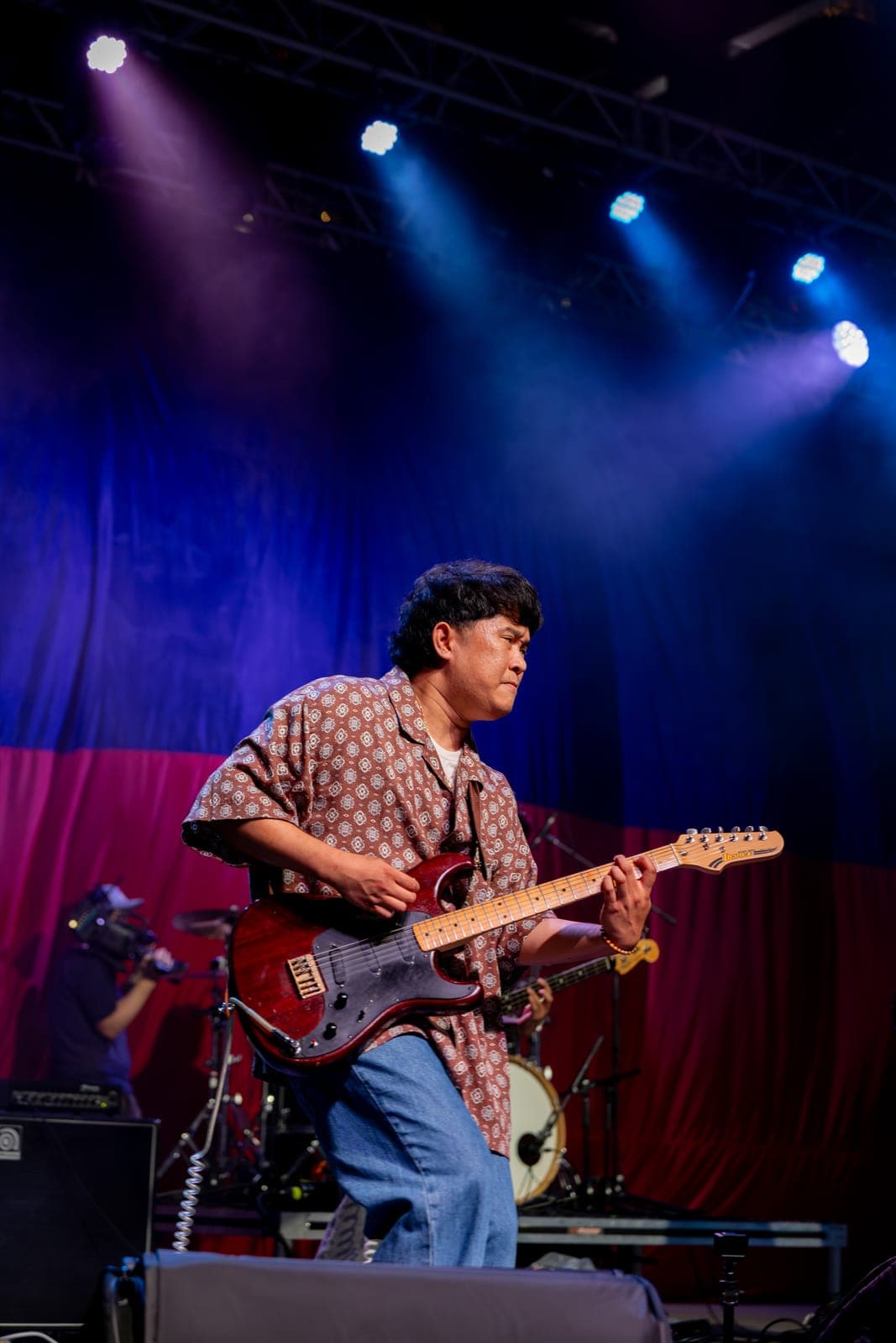 AJ Rafael's guitarist works a red Ibanez in a patterned camp shirt, lit blue against the Philippine flag backdrop