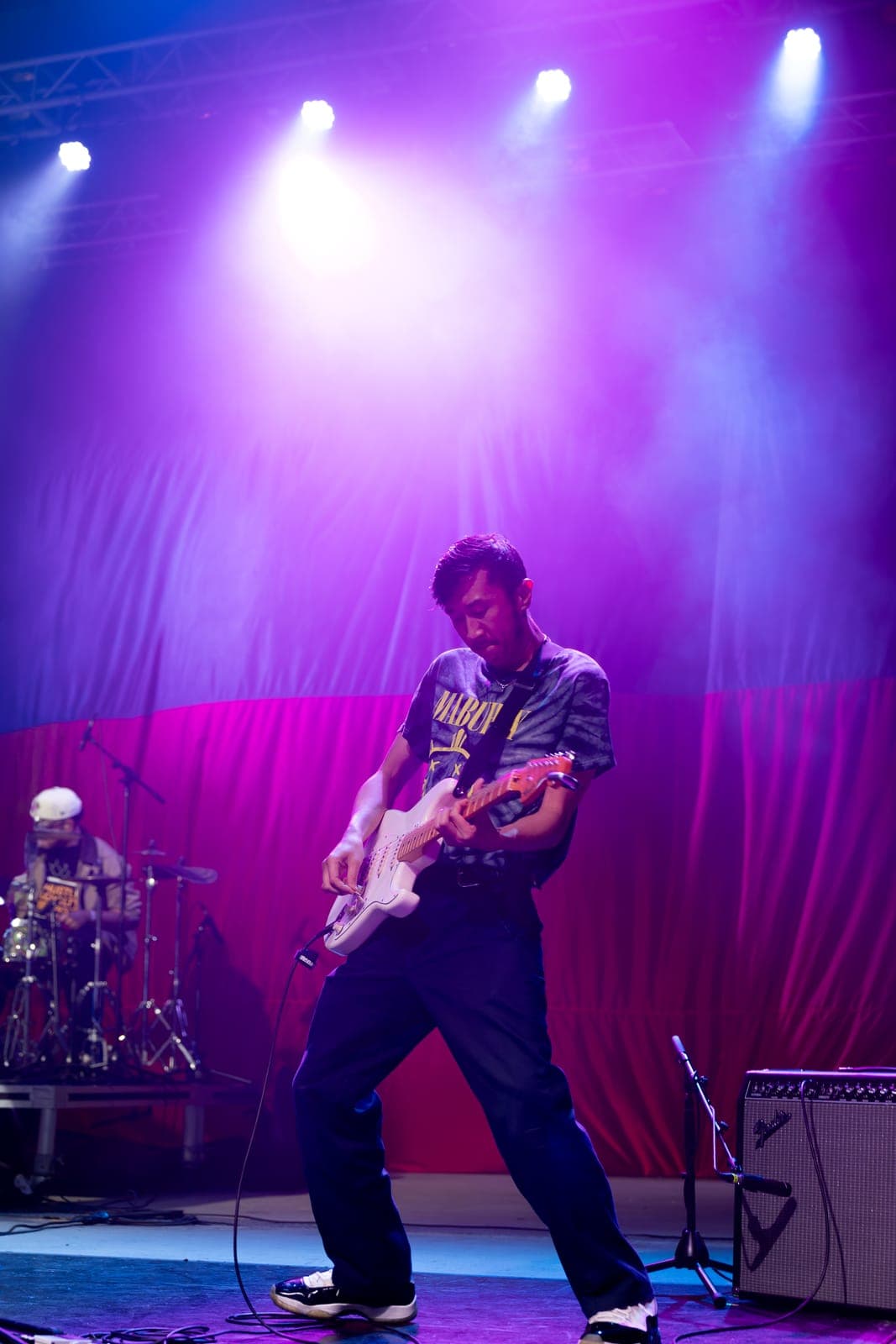 A guitarist in a MABUHAY tie-dye tee digs into a white Stratocaster under magenta light at Philippine Festival Mississauga, the drummer visible behind him