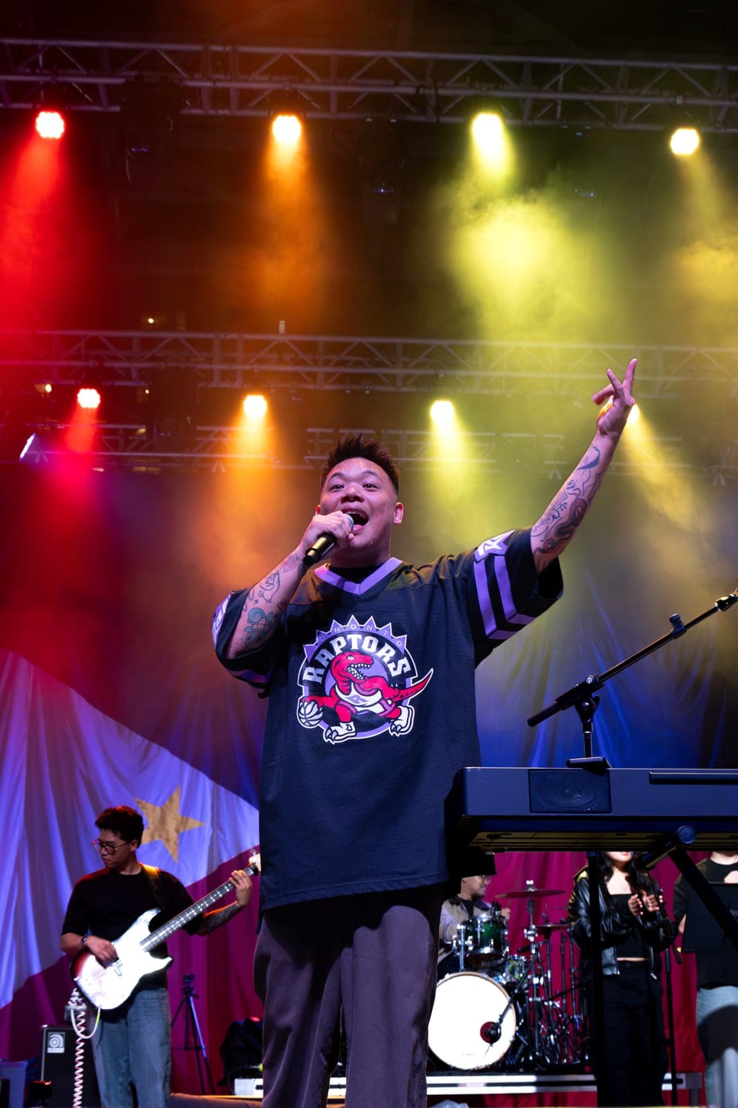 AJ Rafael throws an arm up mid-song in a Raptors jersey at Mississauga Celebration Square, the Philippine flag hanging as a backdrop and his bassist playing to his left