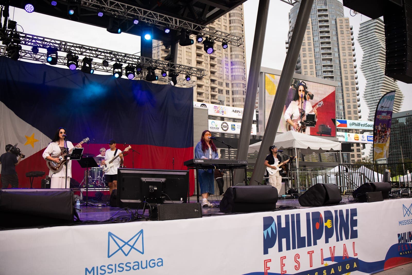A wide view of Simplé's full band on the Celebration Square stage against the Philippine flag, the PHILIPPINE FESTIVAL MISSISSAUGA banner across the barricade below and condo towers rising behind
