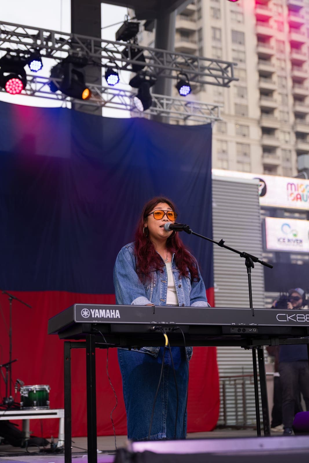 Simplé's vocalist sings into the mic behind a Yamaha CK88 in a denim jacket and orange-tinted glasses, the Philippine flag filling the stage behind her