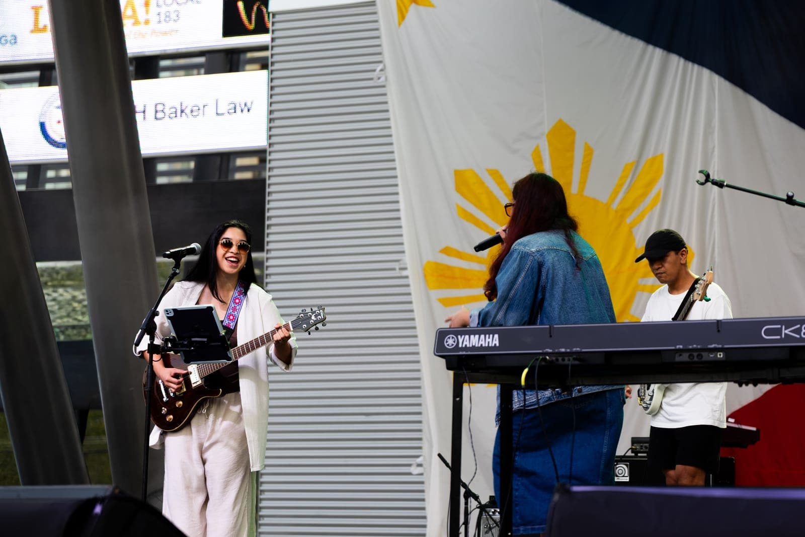 Simplé's guitarist laughs mid-song while the vocalist turns to sing into a handheld mic at her keyboard and the bassist plays on, the Philippine flag's sunburst behind them