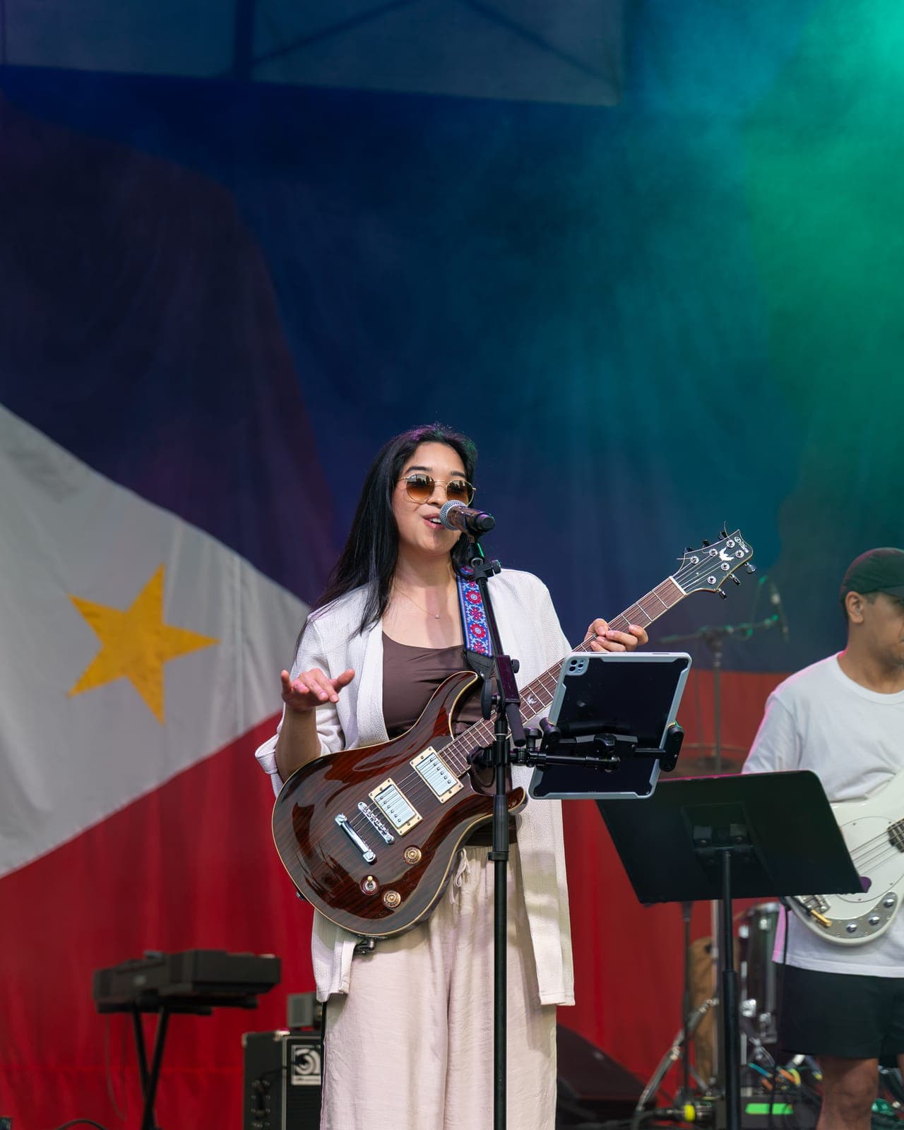 Simplé's guitarist sings into the mic with a dark flame-top guitar under a teal and green wash, an iPad of charts clipped to the stand in front of her