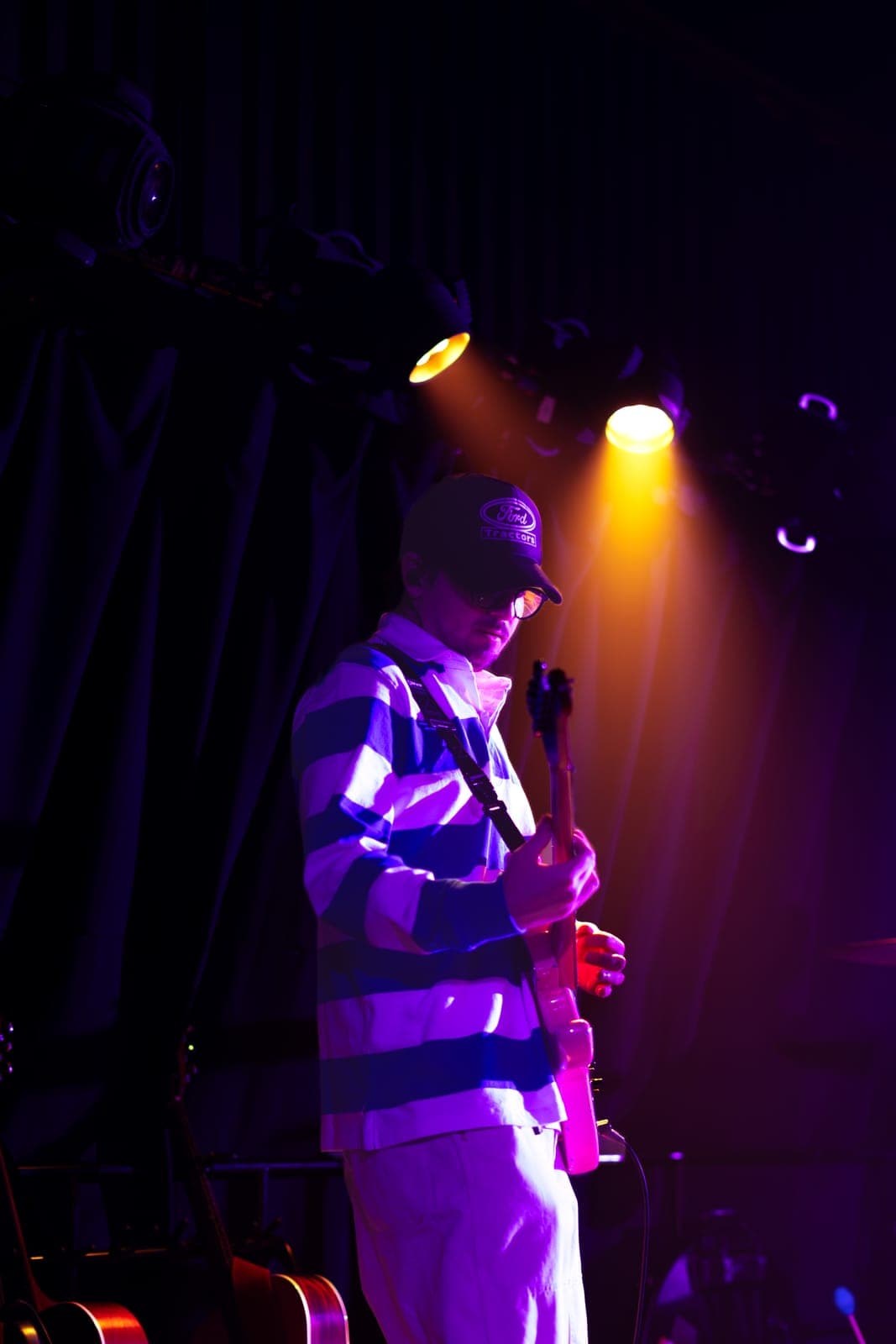 Nia Nadurata's bassist plays under an amber spotlight in a striped shirt and cap at Longboat Hall in Toronto