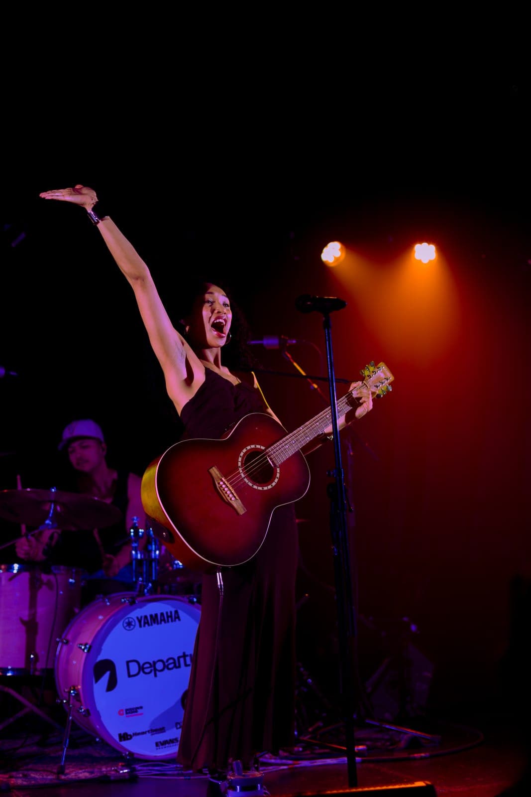 Nia Nadurata raises an arm in triumph while holding an acoustic guitar under warm light at Longboat Hall, Toronto