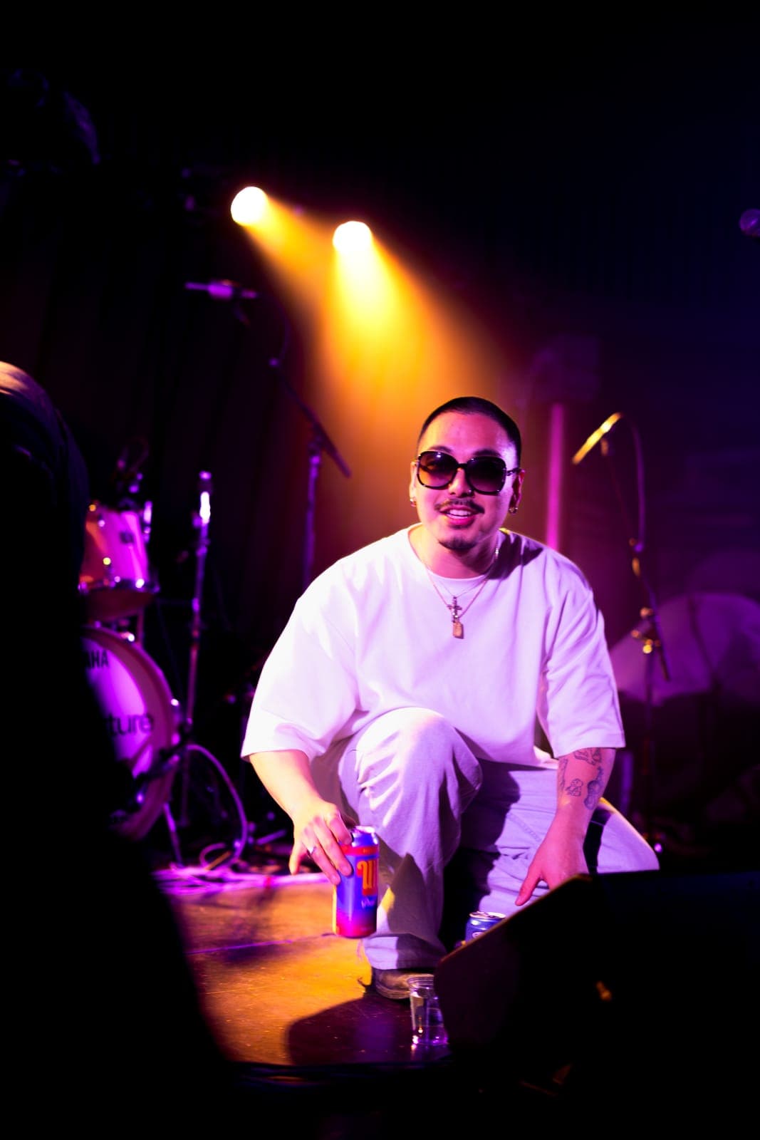 Cherry Kim crouches at the stage edge with a drink, grinning under a golden spotlight at Longboat Hall in Toronto