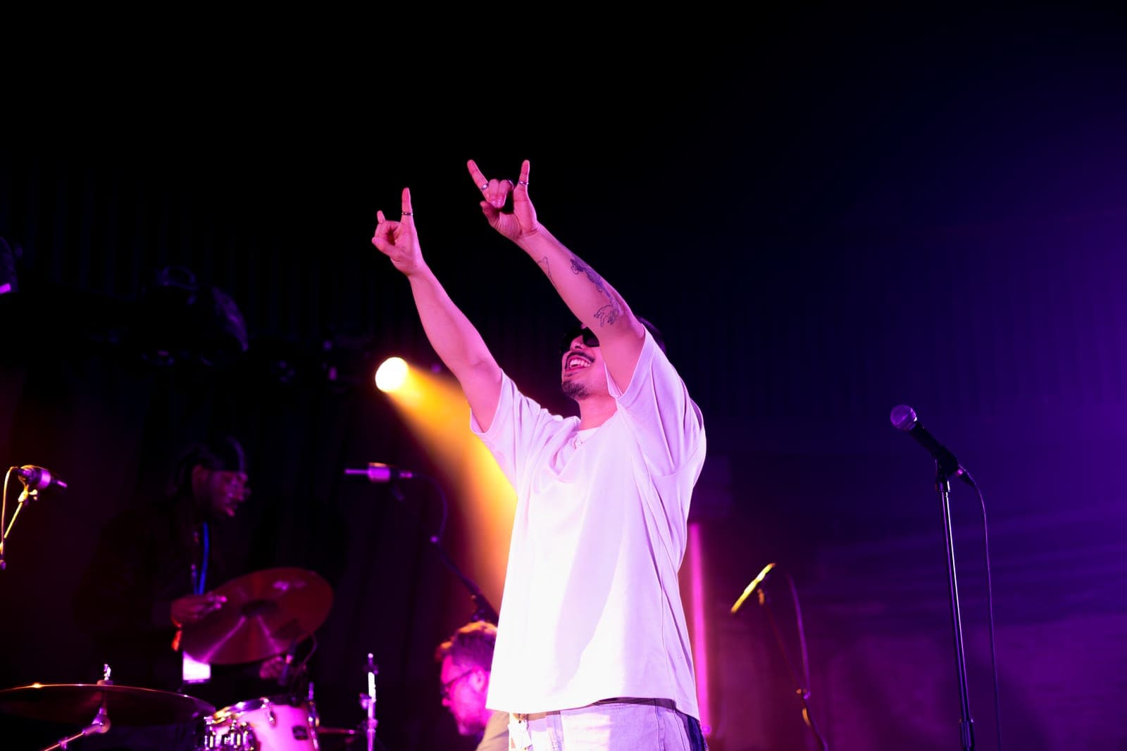 Cherry Kim throws up both hands in rock-on signs, beaming, under purple light at Longboat Hall, Toronto