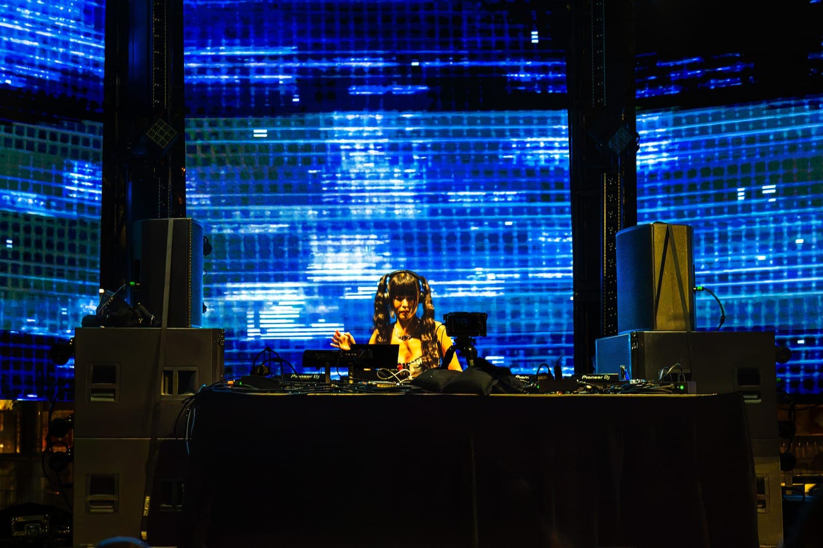 Youna works the Pioneer DJ setup in front of a shimmering blue pixelated video wall, flanked by tall stage speakers.