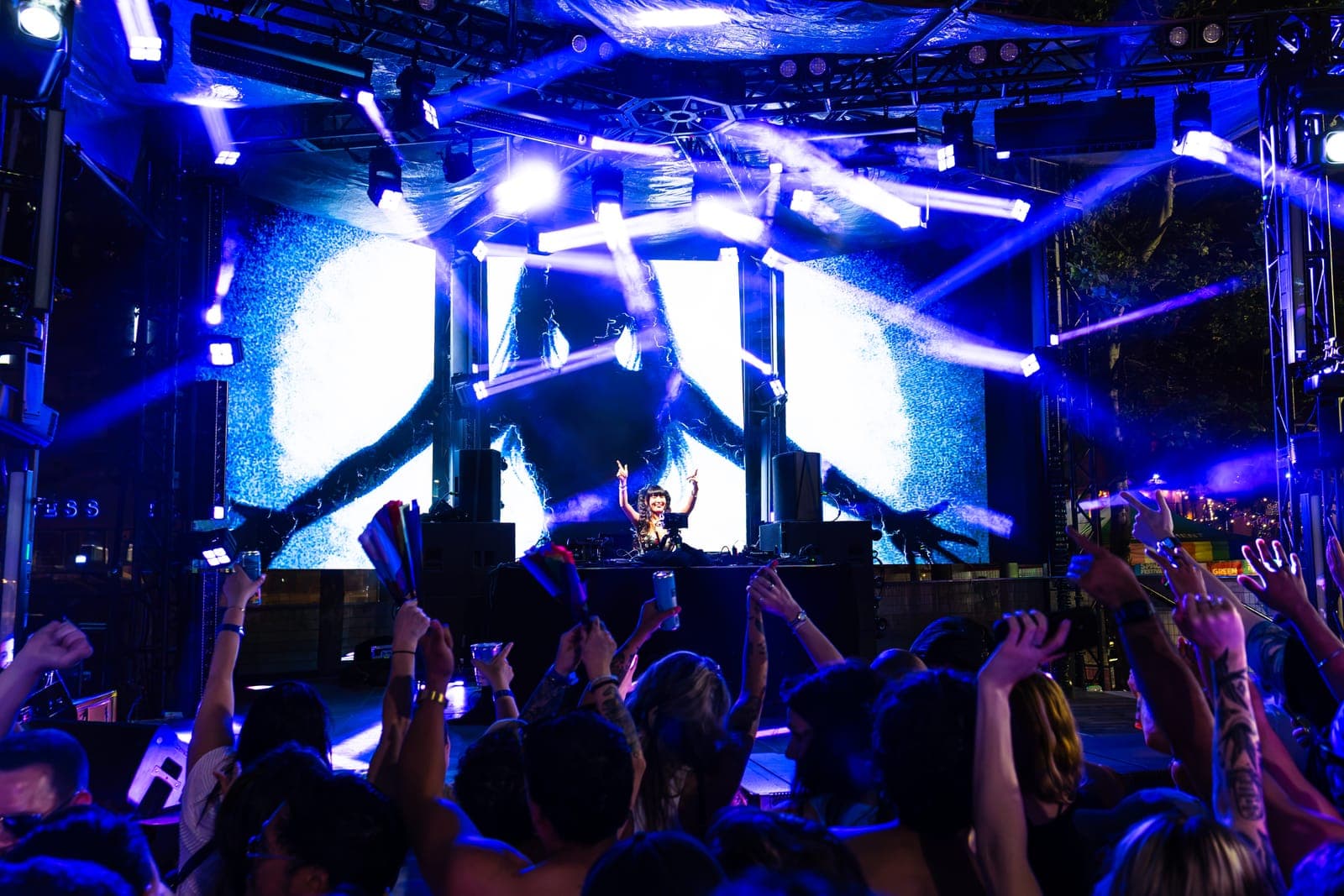 Youna raises her hands over the decks while white beams fan out across a blue video wall and the crowd's arms fill the foreground.