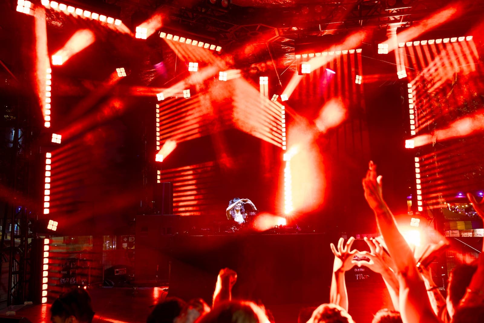 Youna forms a heart with her hands behind the decks amid crossing red light beams, mirrored by a fan making the same heart in the crowd.