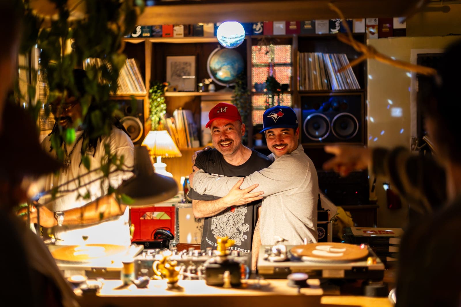 Do Not Push and Kiinjo embrace and laugh behind the decks in front of a wall of vinyl at 915 Dupont in Toronto