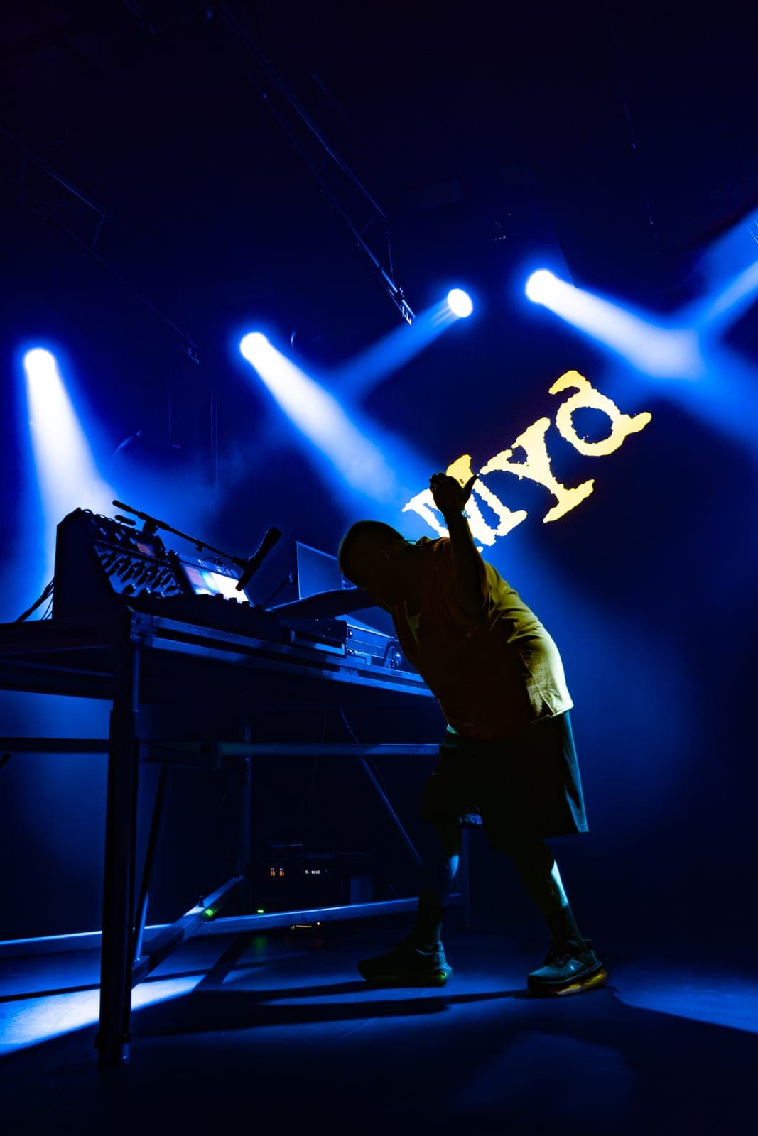 Myd bending down mixing at his gear table with one hand hyping up the crowd beneath the glowing 'myd' logo in blue light at The Mod Club in Toronto