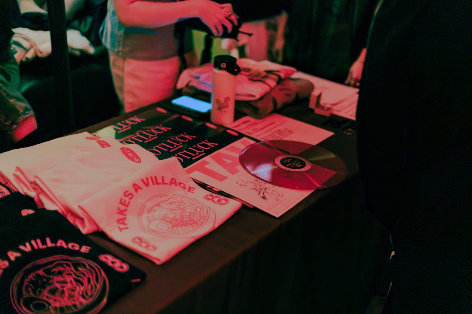 Takes a Village merch tees and a red vinyl of Potluck Volume 1 laid out on the table at The Drake Underground in Toronto