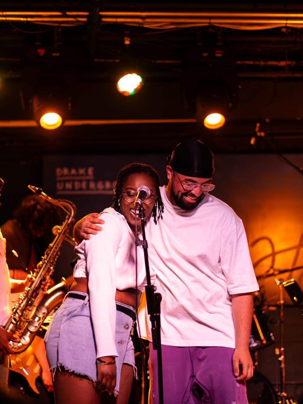 Two Takes a Village members embrace and smile at the mic, the saxophonist beside them, at The Drake Underground in Toronto