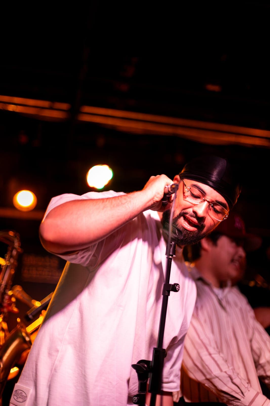 A Takes a Village vocalist sings into the mic in a black cap and glasses under warm light at The Drake Underground in Toronto