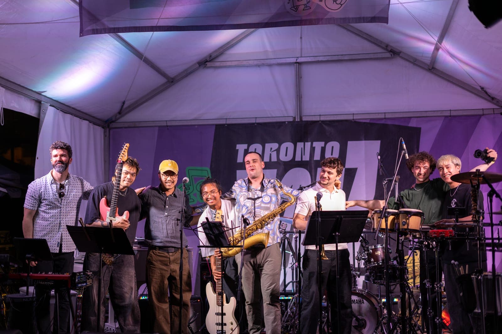 The full Duck Society lineup crowds together grinning for a group photo on the Toronto Jazz Festival stage