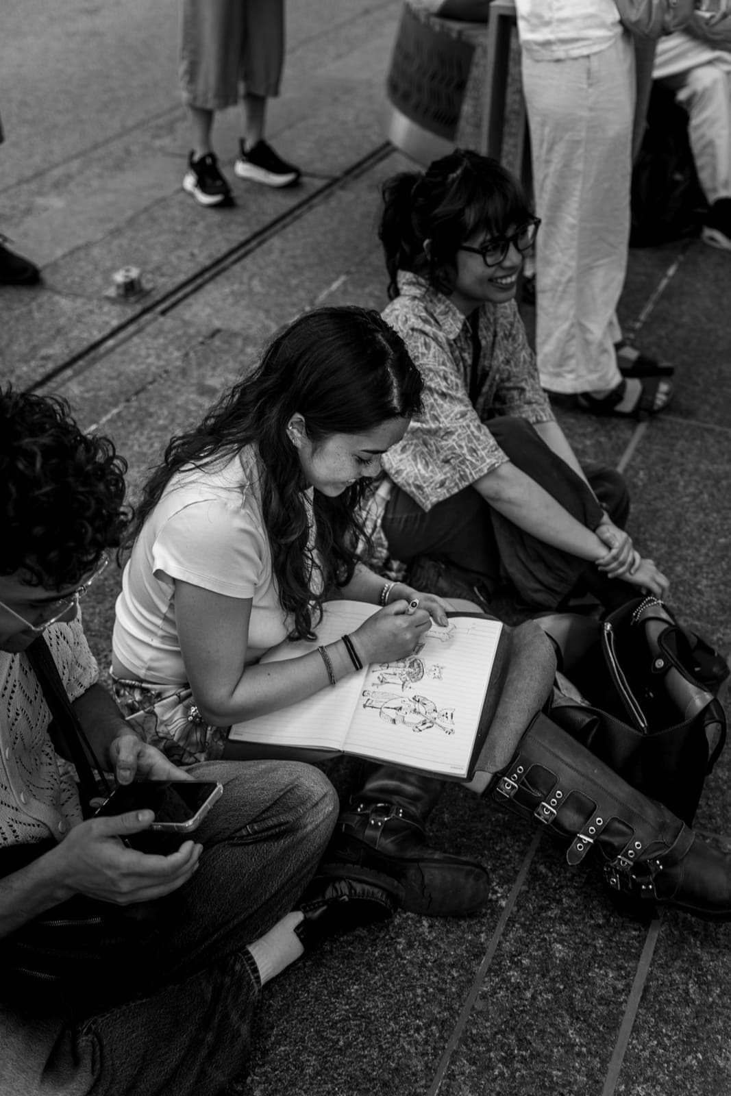 A listener sits on the ground sketching the band in her notebook while a friend beside her smiles