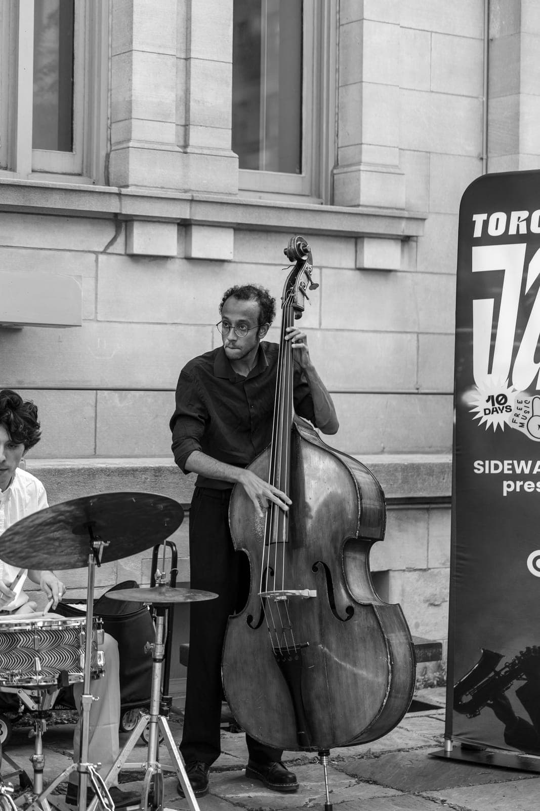 The upright bassist, in glasses and all black, bows a line beside a Toronto Jazz Festival banner