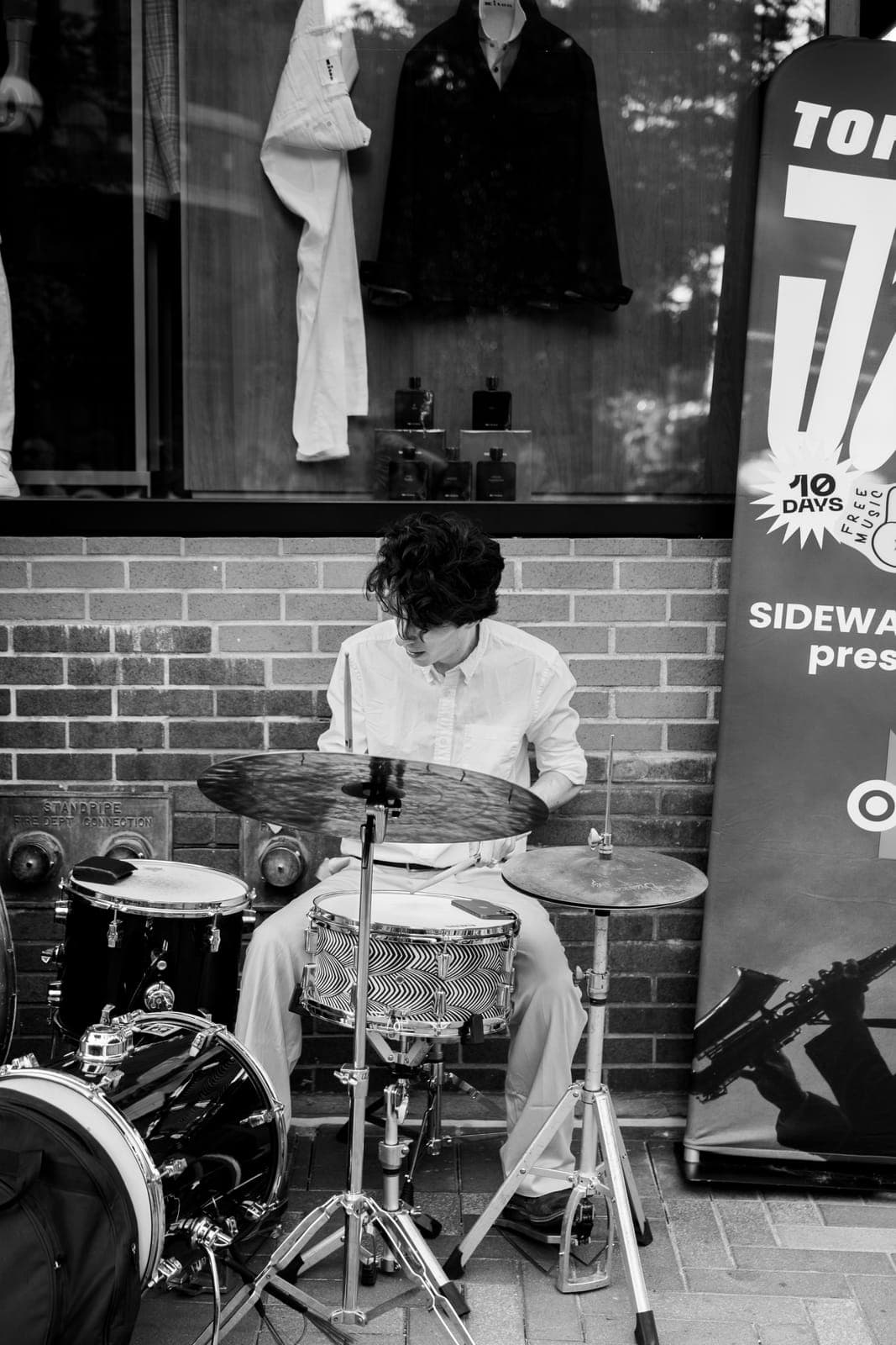 The drummer, in a white shirt, keeps time on a compact kit against a brick storefront wall