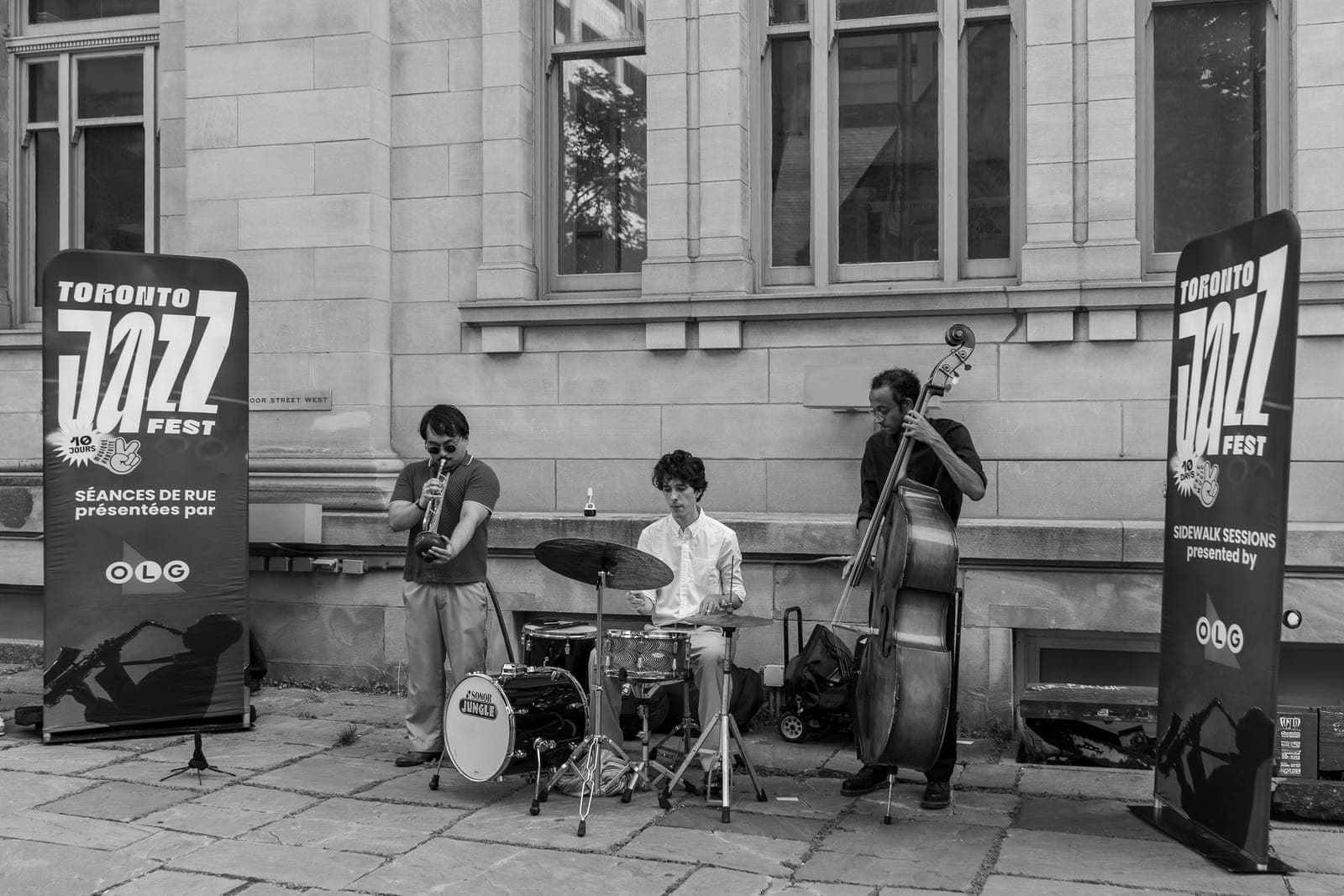 The trumpet, drums, and upright bass trio performs on a stone sidewalk framed by two Toronto Jazz Festival Sidewalk Sessions banners