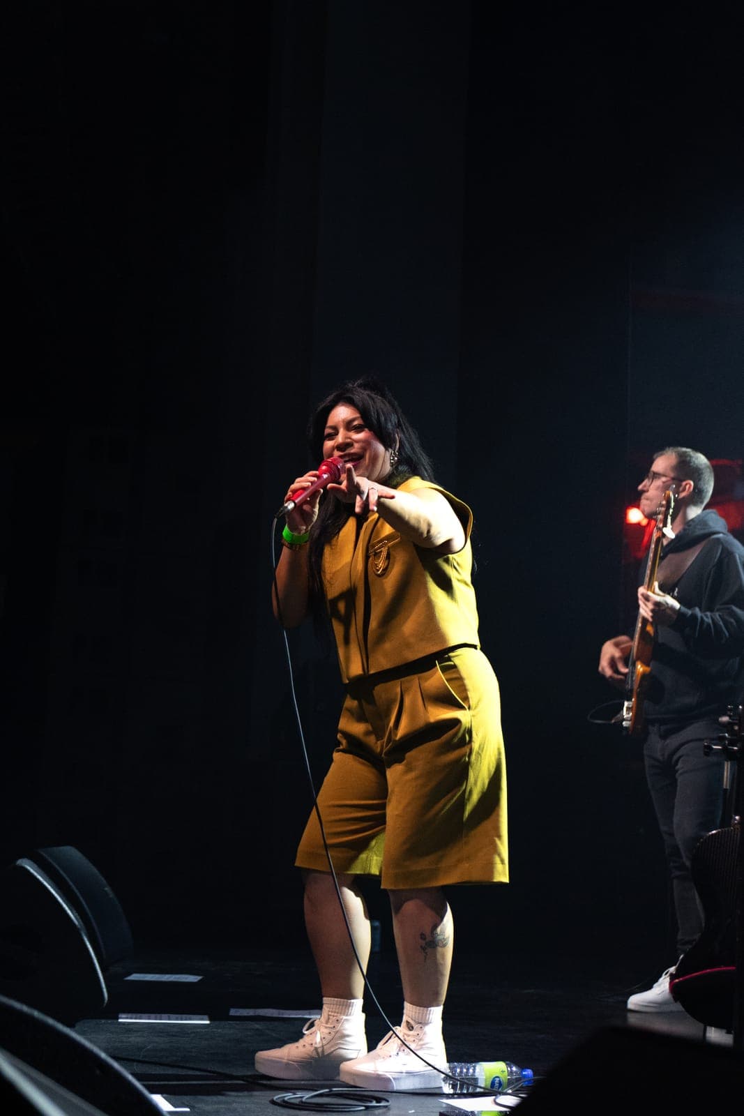 Maya Killtron points to the crowd as she sings into a red microphone in her mustard outfit