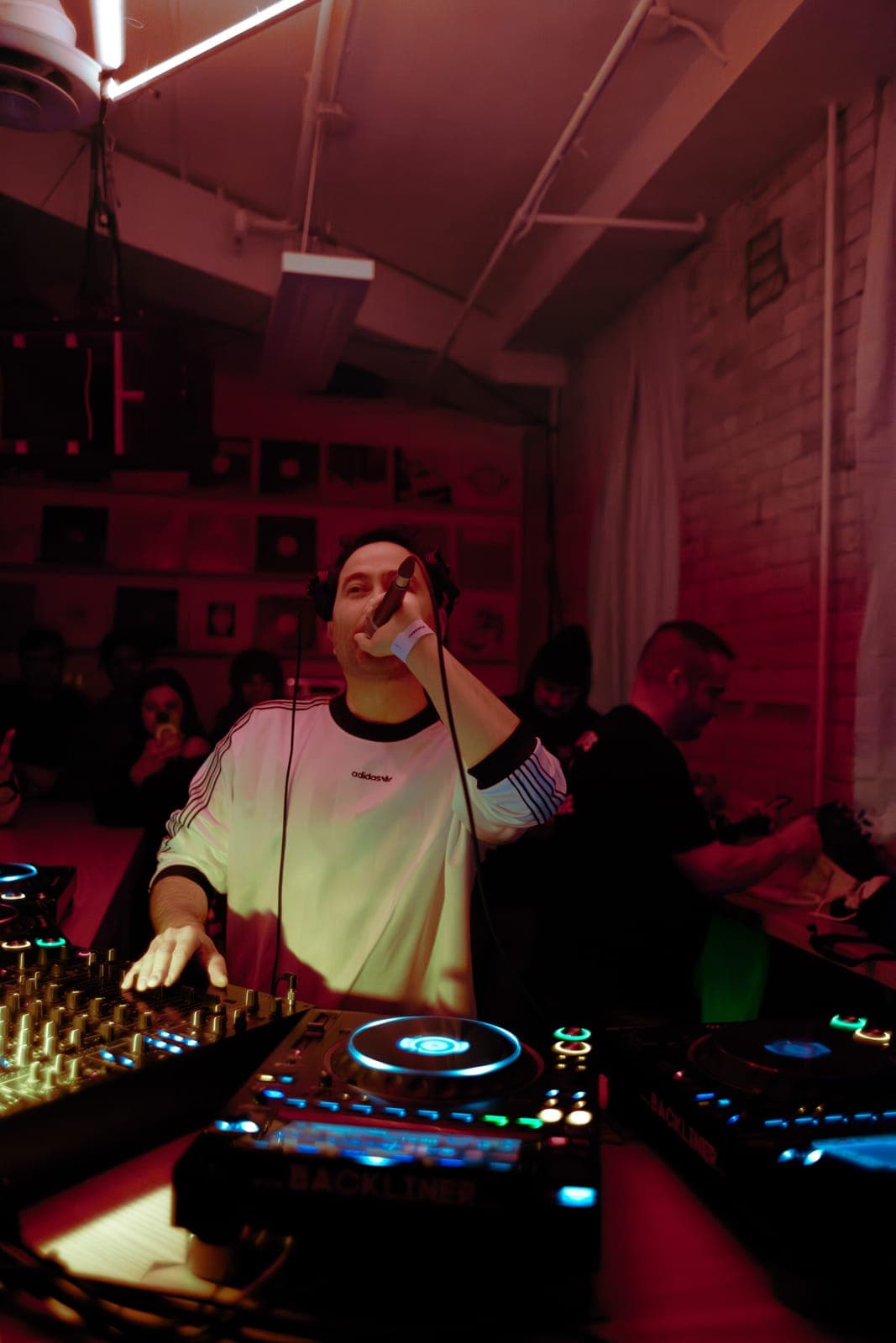 Kiinjo holds the mic and works the decks in an adidas jersey under red light at Rhythm in Toronto